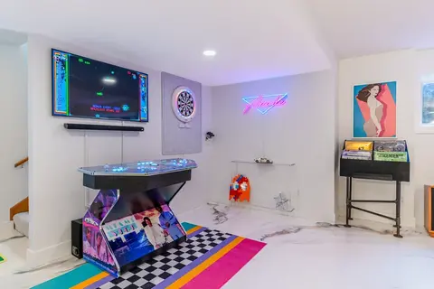 Custom arcade machine in a home game room featuring a wall-mounted TV, dartboard, and retro arcade cabinets